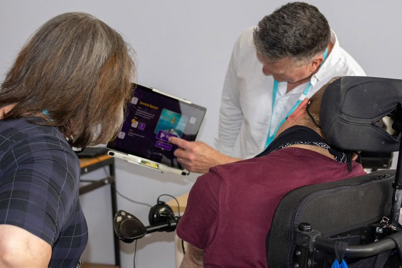 Two occupational therapists work alongside a wheelchair user during an eye gaze communication assessment, demonstrating a tablet-based AAC system mounted for accessible communication.