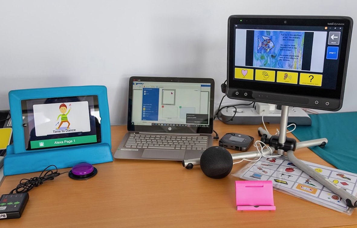 A selection of assistive technology equipment displayed on a table, including speech-generating devices, AAC software, switches, symbol communication boards, and access tools used to support communication and participation.