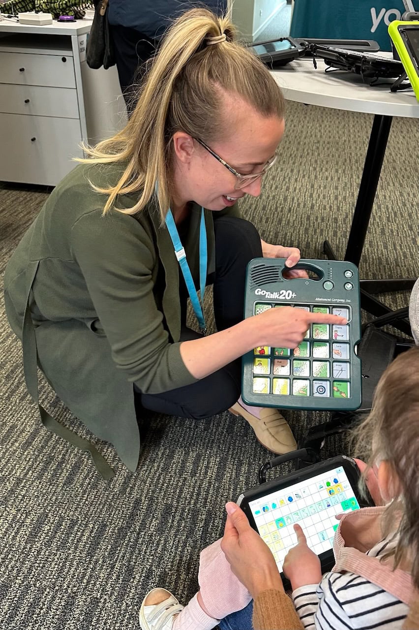 A speech pathologist demonstrates a GoTalk 20+ communication device to a child who is using a tablet-based AAC system, supporting communication through symbol-based vocabulary and assistive technology.